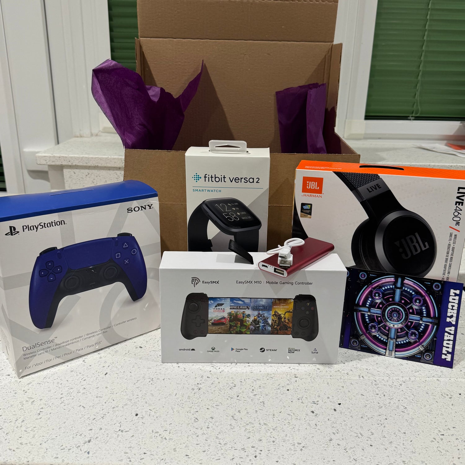 An unboxed mystery box containing a ps5 controller, fitbit, JBL headphones, a phone gaming controller & more.